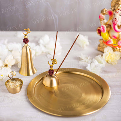 A set of brass pooja items including a bell, a diya, and an incense holder, with rudraksha embellishments, displayed on a white surface with flowers around it.